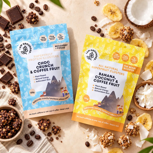 Two stand-up pouches of i am Grounded Organic Superfruit Protein Clusters are arranged on a light beige surface with coffee beans, chocolate pieces, dried banana slices, coconut, and granola around them. The left pouch is light blue and labeled Choc Crunch & Coffee Fruit.