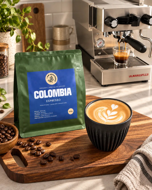 A bag of Colombia Espresso coffee beans by Tricolor Coffee sits on a wooden cutting board next to a cup of latte art coffee and an espresso machine in a kitchen setting.