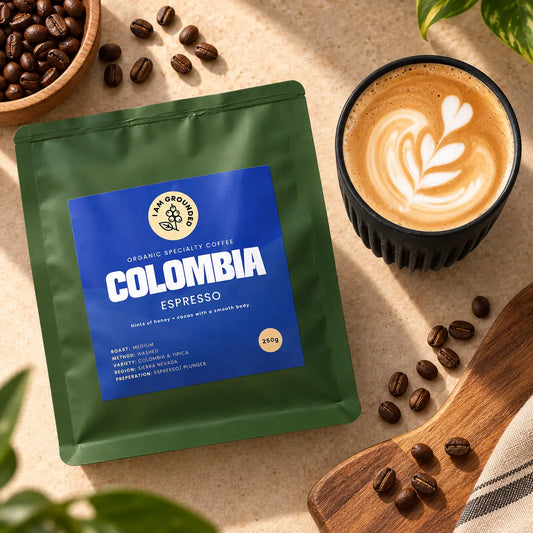 A green stand-up coffee bag labeled  Colombia Espresso' is arranged on a beige surface beside a cup of latte art coffee, with scattered coffee beans and a wooden spoon in the background.