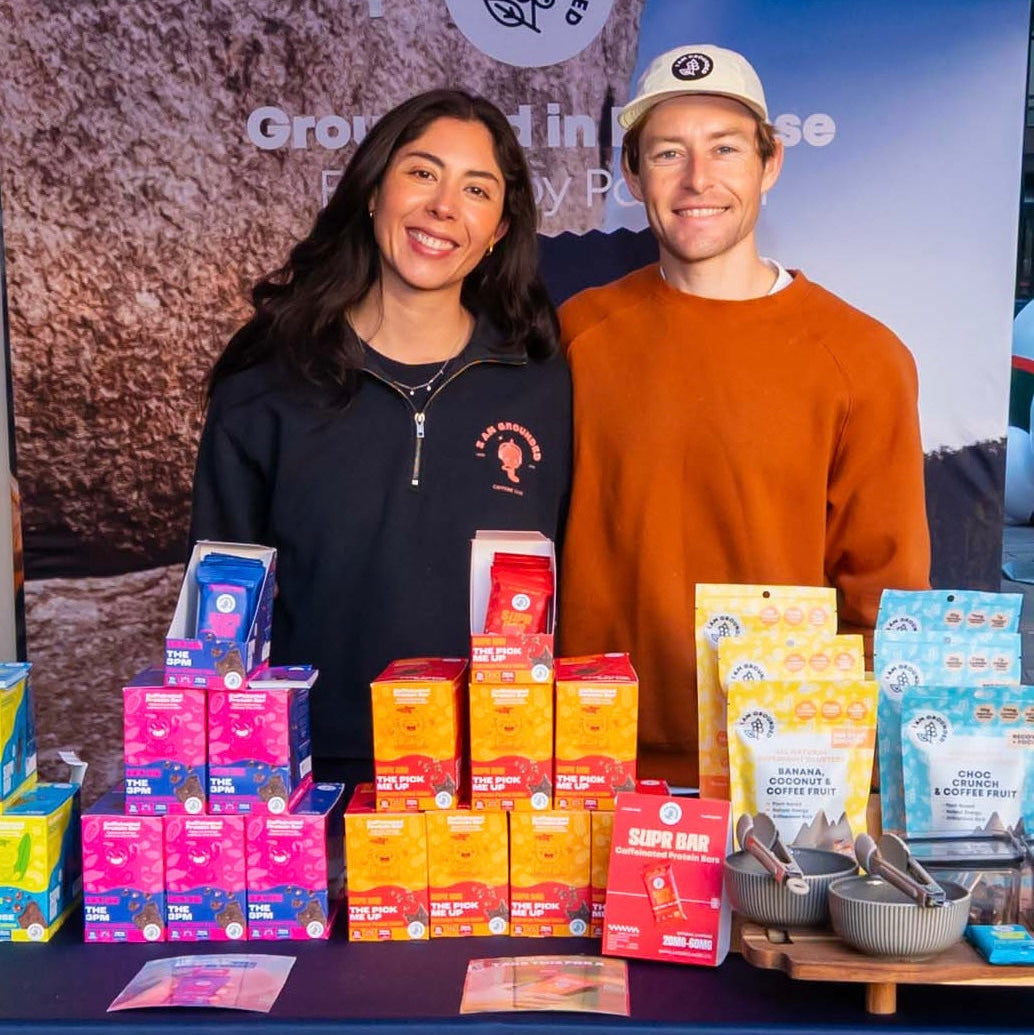 A man and a woman stand behind a table displaying various colorful packaged tea products and bowls on a blue tablecloth at an outdoor event. The man wears an orange sweatshirt and a white cap, and the woman wears a dark navy quarter-zip pullover.