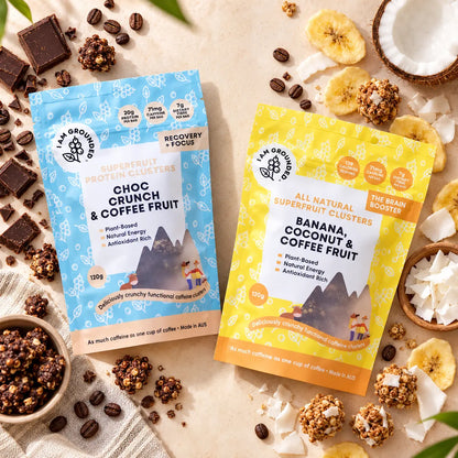 Two stand-up pouches of i am Grounded Organic Superfruit Protein Clusters are arranged on a light beige surface with coffee beans, chocolate pieces, dried banana slices, coconut, and granola around them. The left pouch is light blue and labeled Choc Crunch & Coffee Fruit.