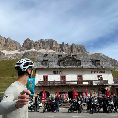Person holding a product in front of a mountain lodge with people and motorcycles in the background