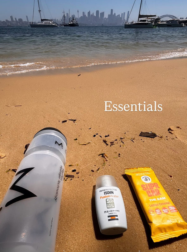 Three beach essentials including a water bottle, sunscreen, and towel on a sandy beach with a city skyline in the background.